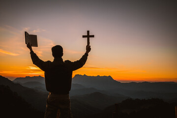 The silhouette of a young man holding a holy bible and raising a christian cross religious symbol...
