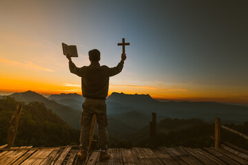 The silhouette of a young man holding a holy bible and raising a christian cross religious symbol...