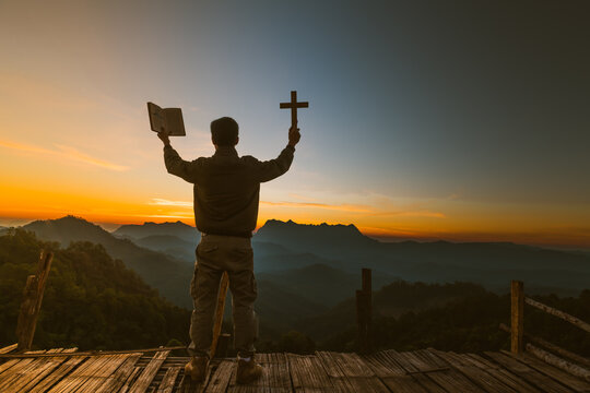 The silhouette of a young man holding a holy bible and raising a christian cross religious symbol in the light and scenery over the sunrise religious background faith concept