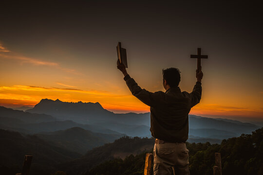 The silhouette of a young man holding a holy bible and raising a christian cross religious symbol in the light and scenery over the sunrise religious background faith concept
