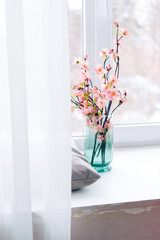 Soft home decor, glass jug, vase with white and pink beautiful flowers against window and curtains and table top. interior. 