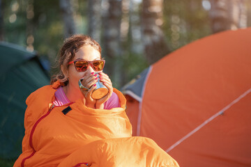 Woman in a sleeping bag drinks tea against the backdrop of many tents in a camping site at early...