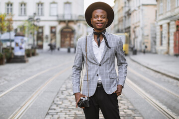 Handsome african man stylish suit and hat posing on city street with retro photo camera in hands. Young male hipster spending weekends for travelling.