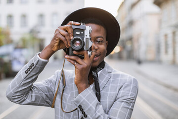 Portrait of young african man wearing trendy hat and suit taking photo on retro camera while walking on street. Concept of people, travelling and lifestyles.