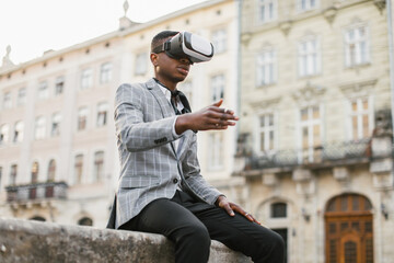 Young african man in stylish suit sitting on city street and wearing VR goggles. Handsome businessman using modern device for work outdoors.