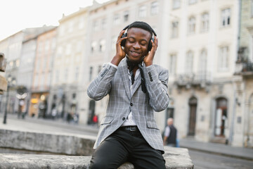 African american man listening music in wireless headphones while resting outdoors. Handsome young guy enjoying favorite song with with closed eyes.