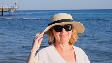 Portrait of a contented 50 year old woman wearing a straw hat and sunglasses enjoying the sun against the blue sea. Summer, vacation, vacation, active retirees