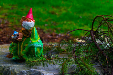 A garden gnome sits on top of a lizard who lays on a rock behind a branch in front of a grass background.
