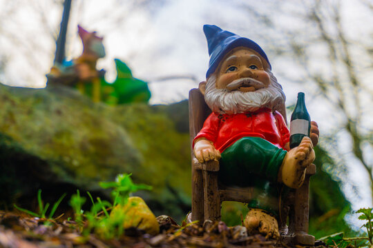 A yard gnome relaxes in his rocking chair with a garden gnome in the backdrop, riding a lizard looking on.