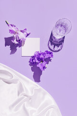 Glass with water and card note with iris flower on pastel violet background with white silk cloth. Summer refreshment concept. Sunlit flat lay. Minimal style. Top view