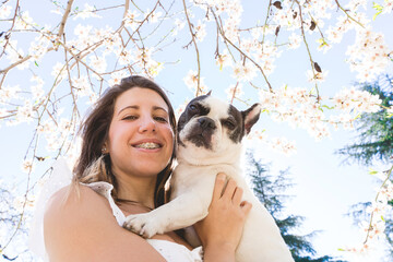 Low angle portrait of woman hugging her dog under almond flowers leaves in springtime. Horizontal portrait of caucasian woman holding french bulldog outdoors. Animals and people lifestyle.