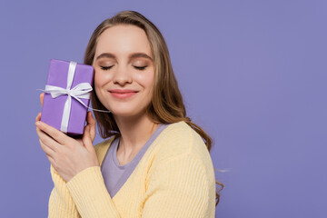 pleased young woman holding wrapped gift box isolated on purple.