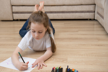 cute little girl in jeans lies on the floor at home and draws with colored pencils, the concept of...