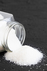 Glass jar with sugar black background.Diabetes. Scattered white sugar on a black table. Copy space. Place for text.Extra calories. Vertical orientation