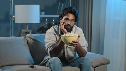 Upset sad bored Arab man tired indian male with beard sits at home evening watching boring TV program switches channel laughing enjoying comedy movie eating popcorn relaxing in dark living room alone