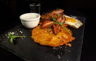 Restaurant serving snacks on the board - potato pancakes with fried meat and egg. A bowl with sauce and a glass of vodka in the background.