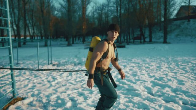 Young muscular man training in a winter park and doing a resistance exercise with a powerlifting band and with a compressed air bottle Drager
