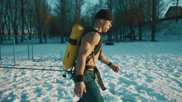 Young muscular man training in a winter park and doing a resistance exercise with a powerlifting band and with a compressed air bottle Drager