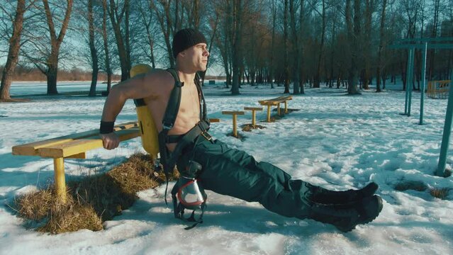 Muscular man with a naked torso and with a drager on his back does reverse push-ups from a bench in a winter park