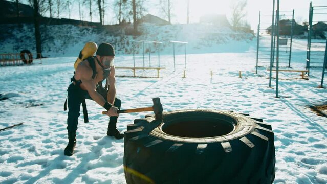 Bodybuilder with a ballon compressed air drager on his back and in a full face mask hits a big tire with a sledgehammer