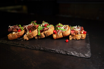 Bruschetta with meat, herbs, and sauce. Restaurant serving on a blackboard on a dark background. A glass of alcohol on the background.
