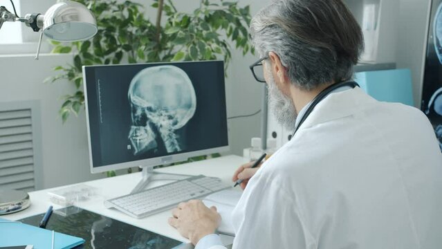 Doctor neurologist looking at human brain MRI images on computer screen and writing in clinic. Hospital routine and modern technology concept.
