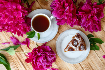 Fototapeta na wymiar Floral tea time with piece of fresh baked berry pie, cup of tea and lush dark pink peony flowers. Morning breakfast with fresh pastry and floral tea. Flat lay, top view.
