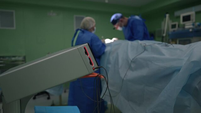 Close-up cautery machine with blurred plastic surgeon and nurse operating patient at background. Caucasian doctor and assistant stitching dissection in slow motion in medical clinic indoors
