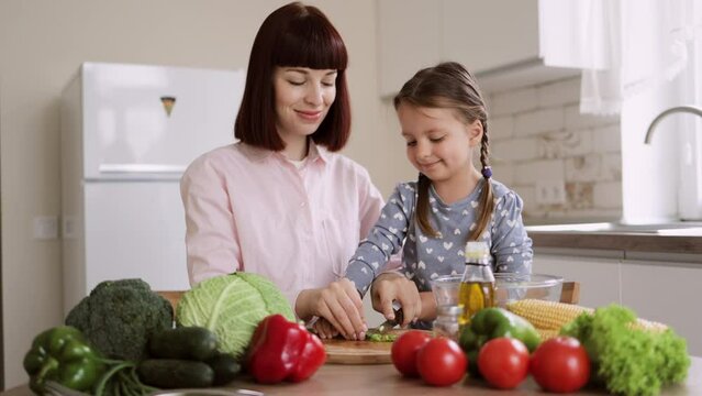 Cute little girl and her beautiful mom in casual clothes are cutting onion and smiling while cooking in kitchen at home. Healthy food at home. Happy family in the kitchen.