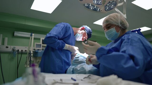 Concentrated doctor cutting stitches with operating scissors as blurred nurse preparing syringe with anesthesia in slow motion. Caucasian man and woman operating patient in surgery room in hospital