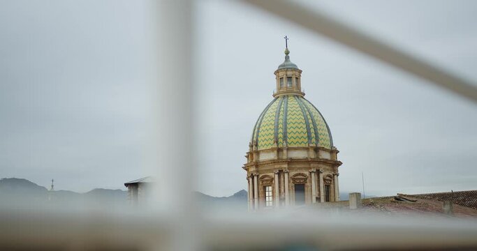 a beautiful green church dome tower with a cross against a cloudy sky and mountains in Palermo, through a blurred foreground
