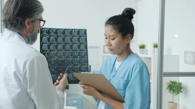 Radiologist Caucasian man examining brain scans while nurse Asian woman writing notes working in hospital together. People and medicine concept.