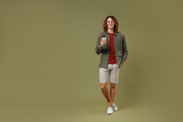 Fototapeta na wymiar Full size body length fun young brunet curly man 20s wear khaki shirt jacket glasses hold takeaway delivery craft paper brown cup coffee to go isolated on plain olive green background studio portrait