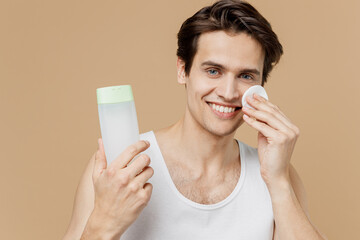 Young smiling man 20s perfect skin in undershirt hold bottle tonic apply moisturizing lotion with cotton pad isolated on pastel pastel beige background Skin care healthcare cosmetic procedures concept © ViDi Studio
