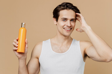 Attractive cool young man 20s perfect skin in undershirt hold bottle of shampoo touch hair isolated...