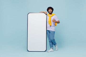 Full size jubilant fun young bearded Indian man 20s wear white t-shirt stand near big mobile cell phone with blank screen workspace area isolated on plain pastel light blue background studio portrait