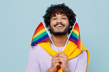 Jubilant happy fancy fun young bearded Indian man 20s years old wears white t-shirt hold raise up to face striped colorful rainbow flags isolated on plain pastel light blue background studio portrait.