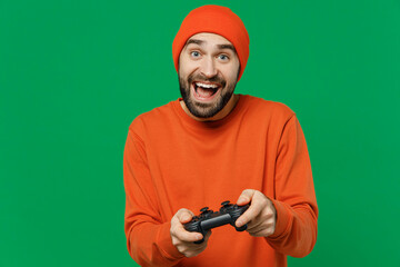 Young overjoyed smiling happy caucasian man 20s wear orange sweatshirt hat hold in hand play pc game with joystick console isolated on plain green background studio portrait. People lifestyle concept.