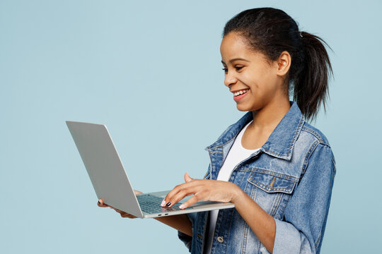 Little Smiling Happy Kid Teen Girl Of African American Ethnicity 12-13 Years Old In Denim Jacket Hold Use Work On Laptop Pc Computer Isolated On Pastel Plain Light Blue Background. Childhood Concept.