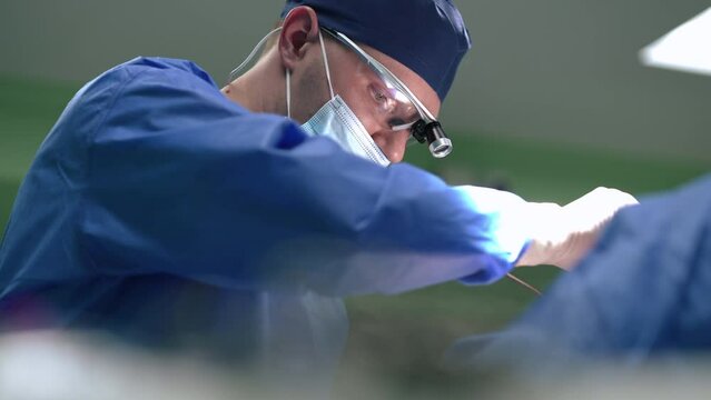 Confident plastic surgeon stitching dissection with cautery machine in slow motion. Side view focused Caucasian man operating patient in surgery room in medical clinic indoors. Technology and medicine