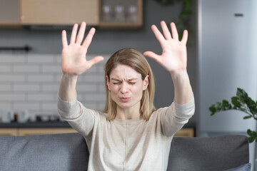 Scared woman sitting on sofa at home, holding hands in front, stop sign