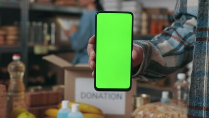 Close up of modern smartphone with green blank screen on my hands. Blur background of woman packing donation boxes. Charitable fund.