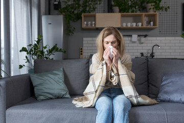 The sick woman is sitting on the couch at home, has a cough and fever, cold and runny nose