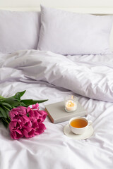 Fototapeta na wymiar Bouquet of pink tulips, cup of tea, burning candle and book on bed with lilac linen.