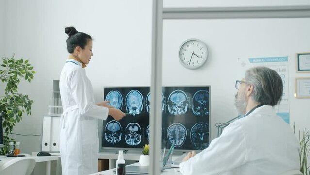 Neurologist Asian lady in white coat discussing MRI images with male colleague Caucasian man talking and pointing at brain scans in hospital