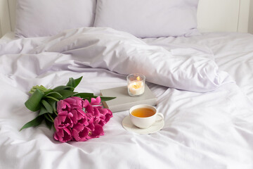 Bouquet of pink tulips, cup of tea, burning candle and book on bed with lilac linen.