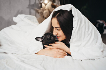 Beautiful brunette woman with a French Bulldog puppy in a bed under a blanket.