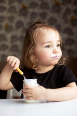 Funny little girl with dirty face eating plain homemade yogurt from glass jar using spoon. Vertical. 18-24 months kid girl in black t-shirt eating yogurt and looking window. Vertical format