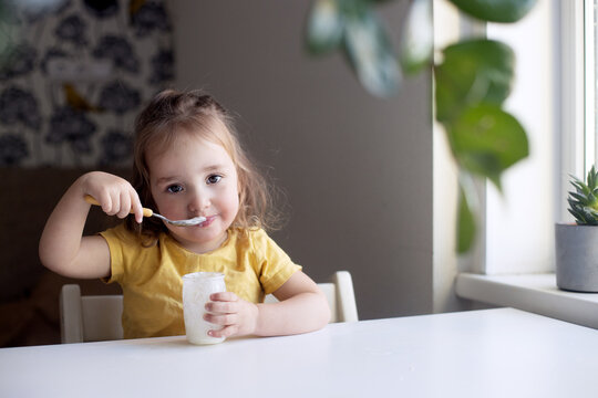 Cheerful Little Girl Eating Plain Homemade Yogurt From Glass Jar Using Spoon. Healthy Smiling Kid, Organic Natural Food. Green Kitchen, Home Plants. Girl 18-24 Months Wearing Yellow R-shirt