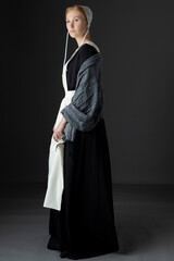 An Amish woman wearing a black dress, white apron, grey shawl, and cap against a grey backdrop